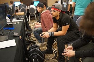 Several people are sitting in a room with gaming consoles and monitors, focusing intently on their screens. The setting appears to be a casual gaming event, with participants holding controllers while playing video games. The room has a casual atmosphere with chairs and tables set up for gaming.