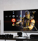 A flat-screen TV displays a video game interface featuring a character from Mortal Kombat in the center. Beside the TV is a stack of video game cases for the PlayStation 3, including titles like FIFA 14 and Grand Theft Auto. Below the TV, there is a gaming console, controllers, and other gaming equipment.