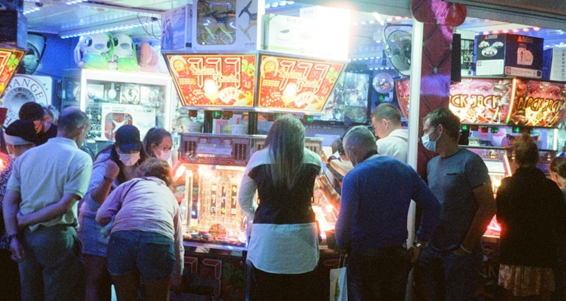 A group of people standing in front of an illuminated amusement arcade, engaging with various gaming machines. The environment is bustling with activity, and colorful lights create a vibrant atmosphere. The ceiling is decorated with large, playful images of dice and other game-related graphics, enhancing the lively mood.