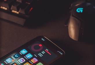 A smartphone displaying various social media app icons is placed on a dark surface. Nearby, a gaming mouse with a glowing logo and a mechanical keyboard with illuminated keys are visible. The lighting is low and focused, creating a moody atmosphere.