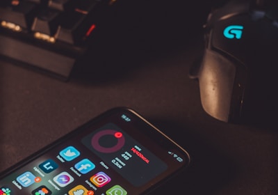 A smartphone displaying various social media app icons is placed on a dark surface. Nearby, a gaming mouse with a glowing logo and a mechanical keyboard with illuminated keys are visible. The lighting is low and focused, creating a moody atmosphere.