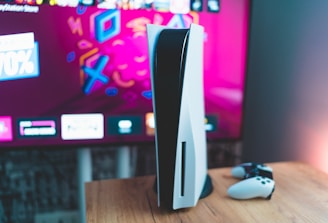 A gaming console is placed on a wooden table with a game controller nearby. In the background, a television screen displays colorful graphics, including geometric shapes and vibrant colors.