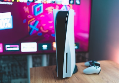 A gaming console is placed on a wooden table with a game controller nearby. In the background, a television screen displays colorful graphics, including geometric shapes and vibrant colors.