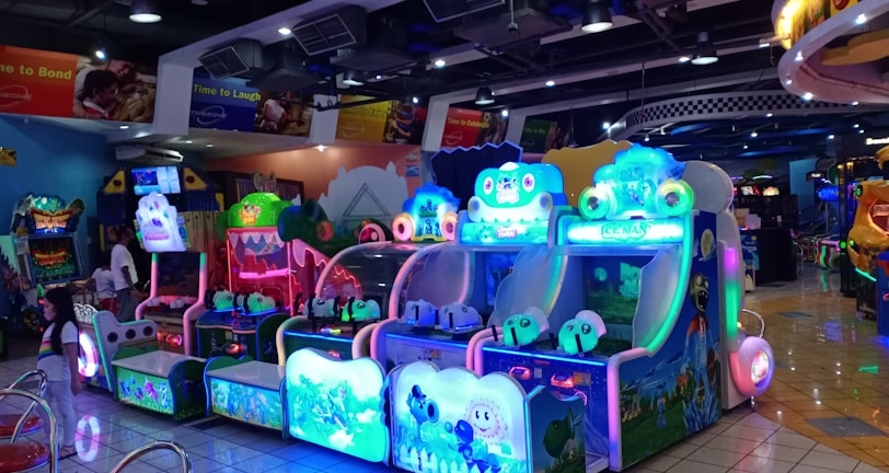 A brightly lit arcade filled with colorful gaming machines sits under ceiling lights. Neon lights illuminate various game stations, creating a playful and vibrant environment. A few red chairs are scattered around the reflective, tiled floor, and a person is observing one of the machines. The atmosphere is lively and cheerful, featuring games with cartoonish designs and engaging themes.